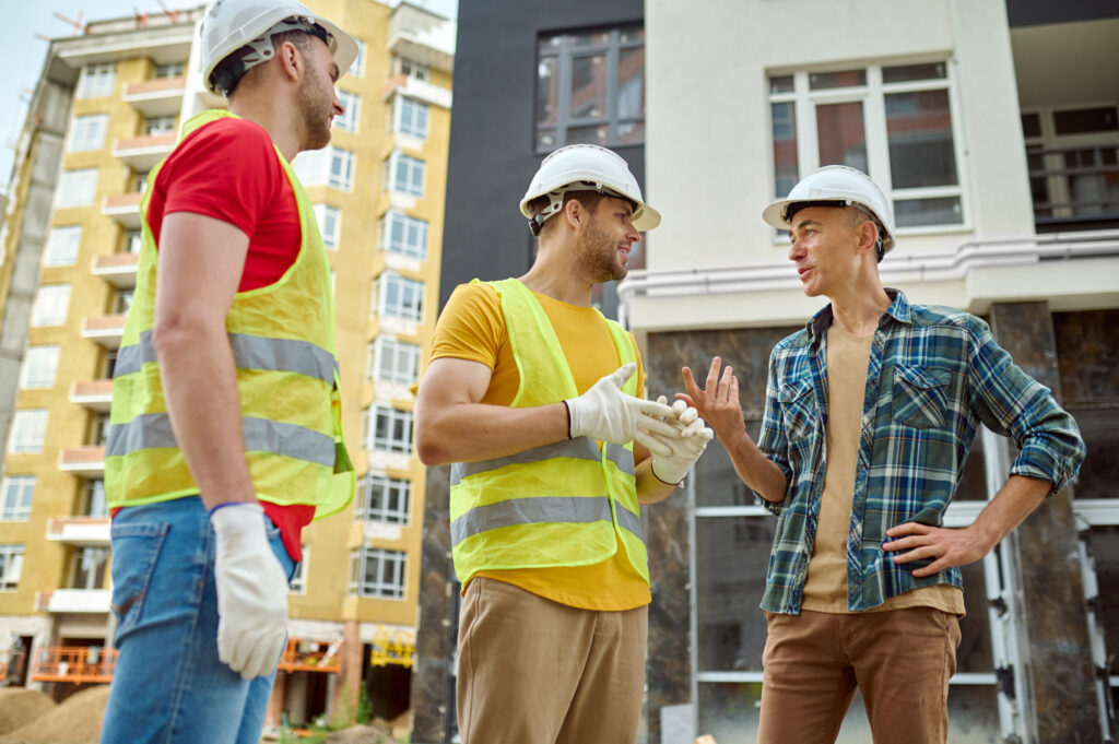 Communication. Engineer in safety helmet and plaid shirt smiling gesture and rude and two workers in gloves and vests listening at construction site during day